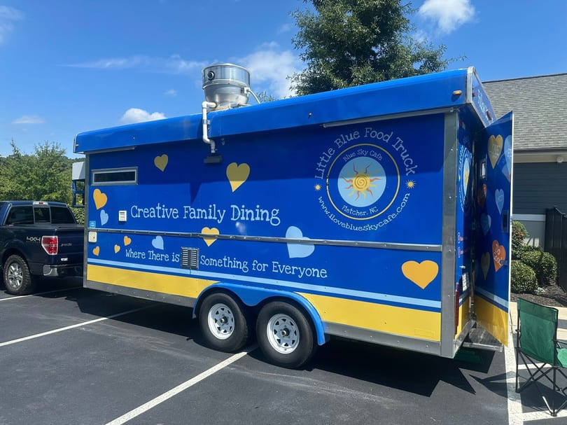 The Little Blue Food Truck, painted blue and yellow with hearts, parked on asphalt.