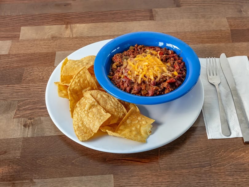 A blue bowl of chili with melted cheese and a side of tortilla chips on a white plate.