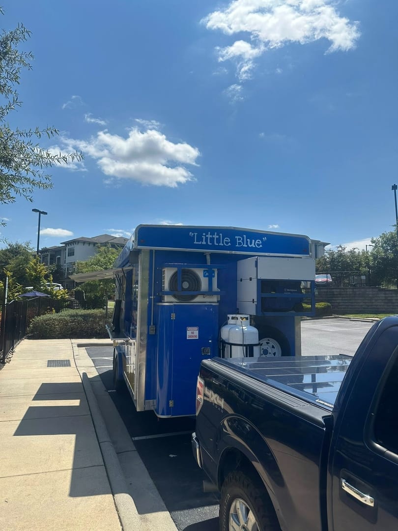 A blue food trailer labeled "Little Blue" is parked next to a black pickup truck on a sunny day.