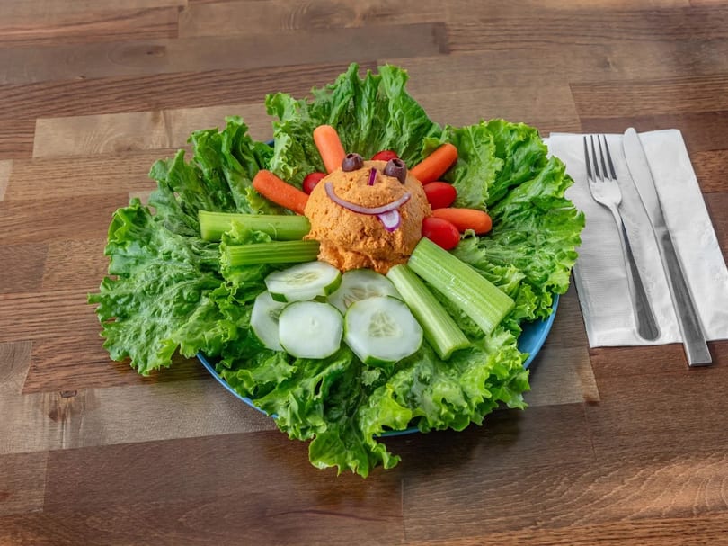 A playful hummus platter shaped like a face with vegetable dippers on a bed of lettuce.